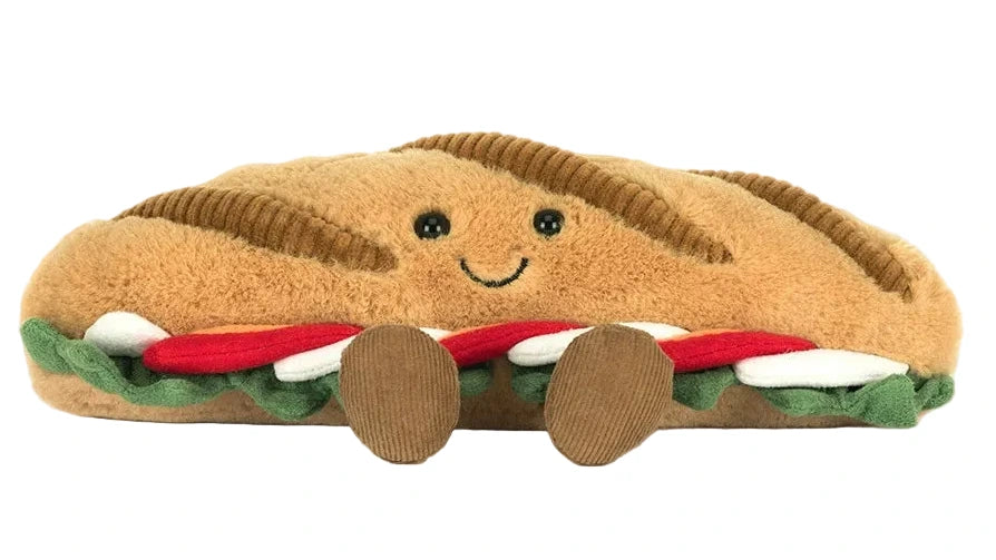 The Jellycat Amuseables Caprese Baguette is a plush toy sandwich with a smiling face, brown bread, green lettuce, red and white layers, and two cute brown feet.