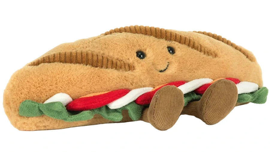 The Amuseables Caprese Baguette by Jellycat is a plush toy shaped like a sandwich, featuring a smiling face, lettuce, tomato, and two round legs—adorably delightful for all ages.