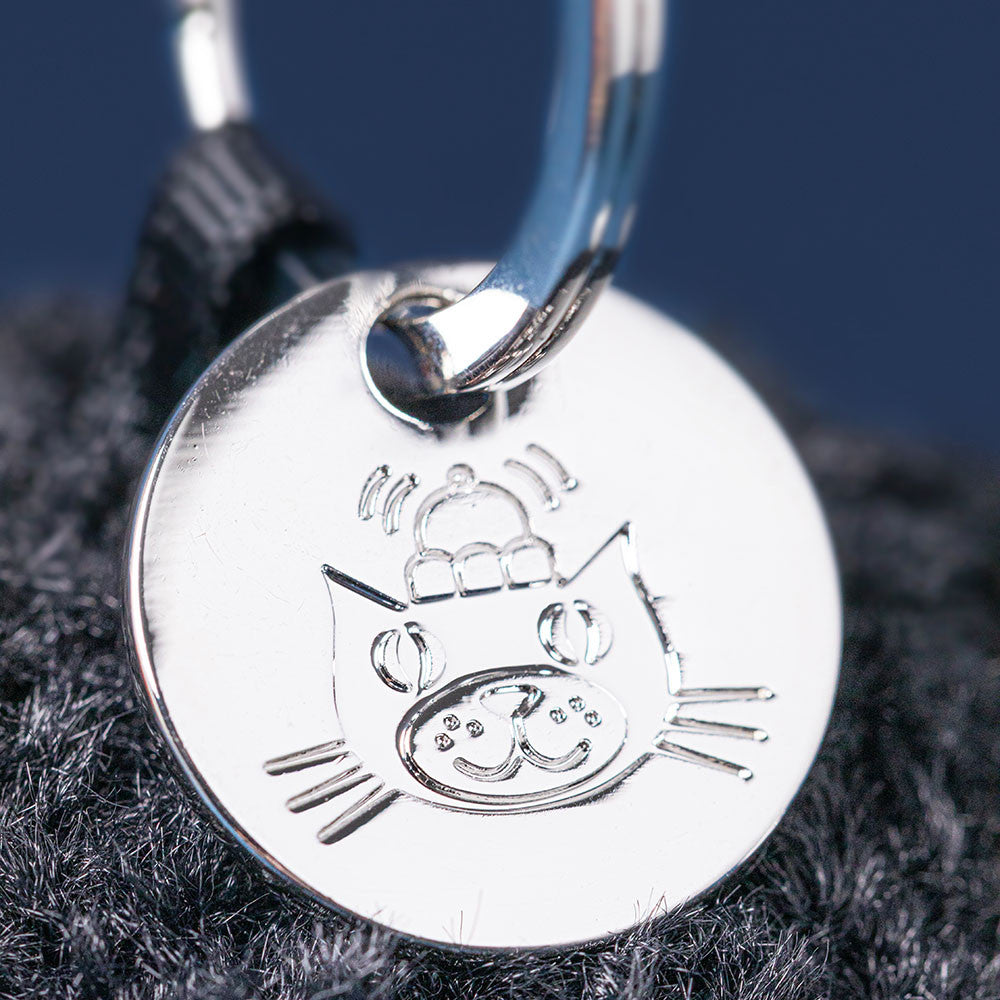 Close-up of the Jellycat Amuseables Black Hole Bag Charm—a round silver tag engraved with a cartoon cat in a hat, attached to a metal ring. Perfect as a quirky physics gift or unique accessory for Jellycat fans.