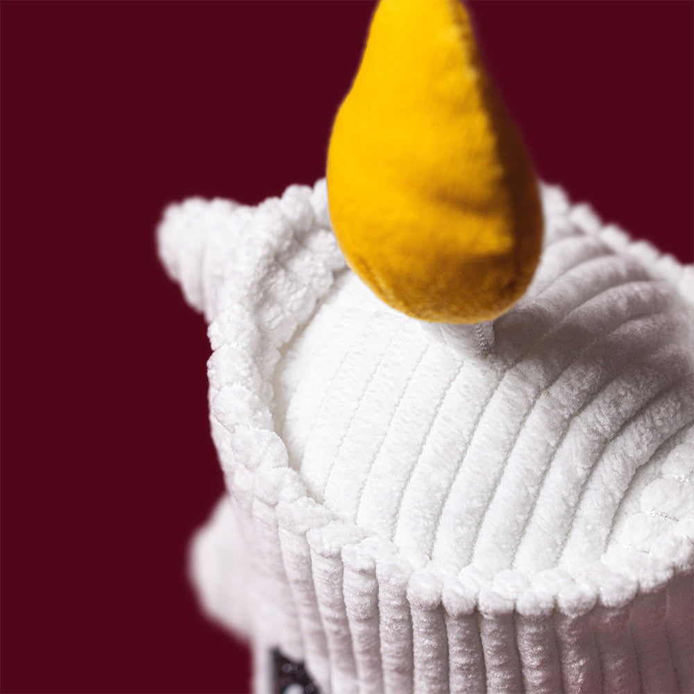 A close-up of the Jellycat Amuseables Flickerty Candle plush toy, featuring soft white textured fabric and a pointed yellow flame on top, set against a solid burgundy background. An ideal whimsical decor piece for any Halloween party planner.