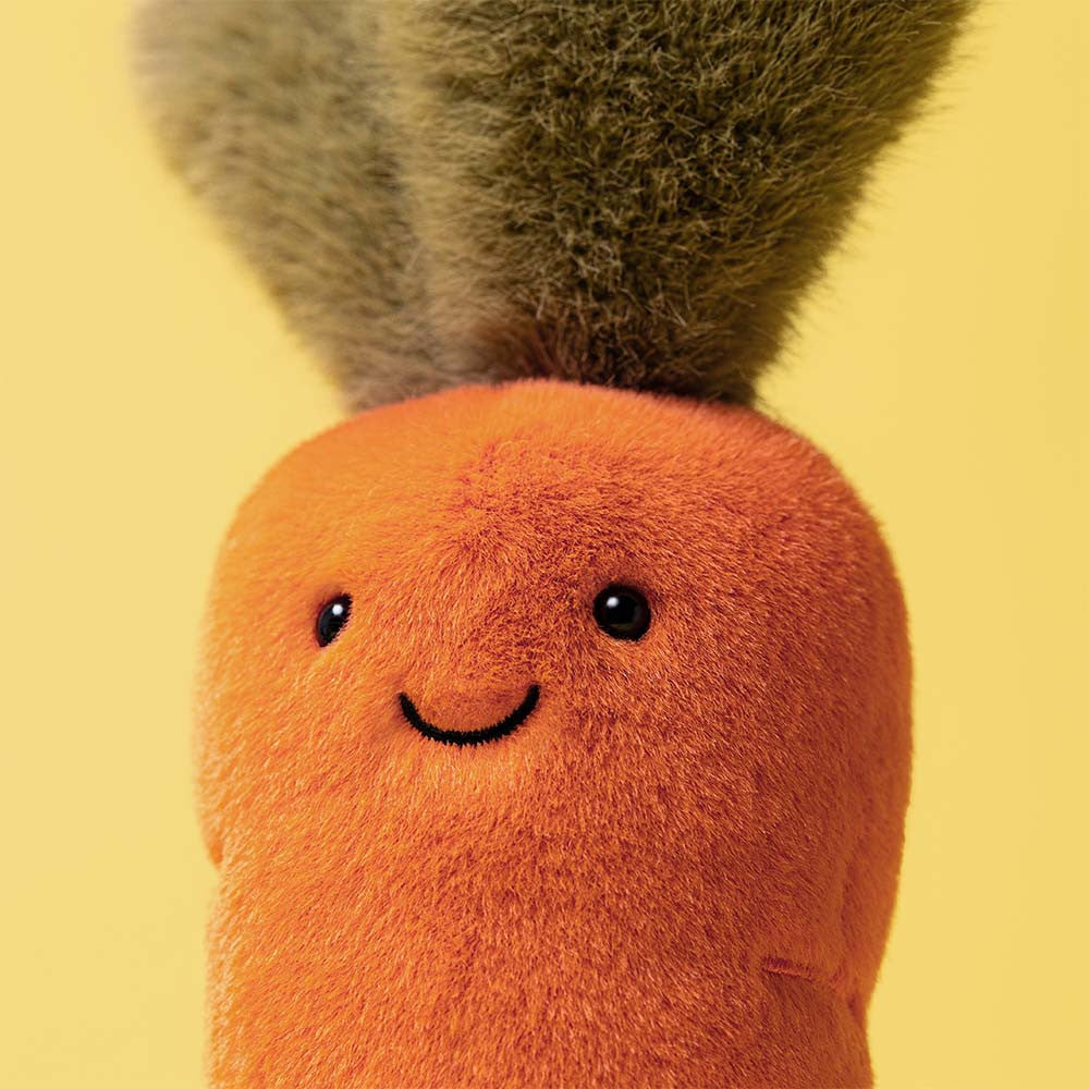 Close-up of the Jellycat Amuseables Carrot plush, featuring a cheerful face, black eyes, and a green leafy top against a yellow background.