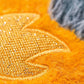 Close-up of the Jellycat Amuseables Space Comet plush toy, featuring textured yellow fabric with gold embroidery and orange fur, next to a patch of gray and black fuzzy material.