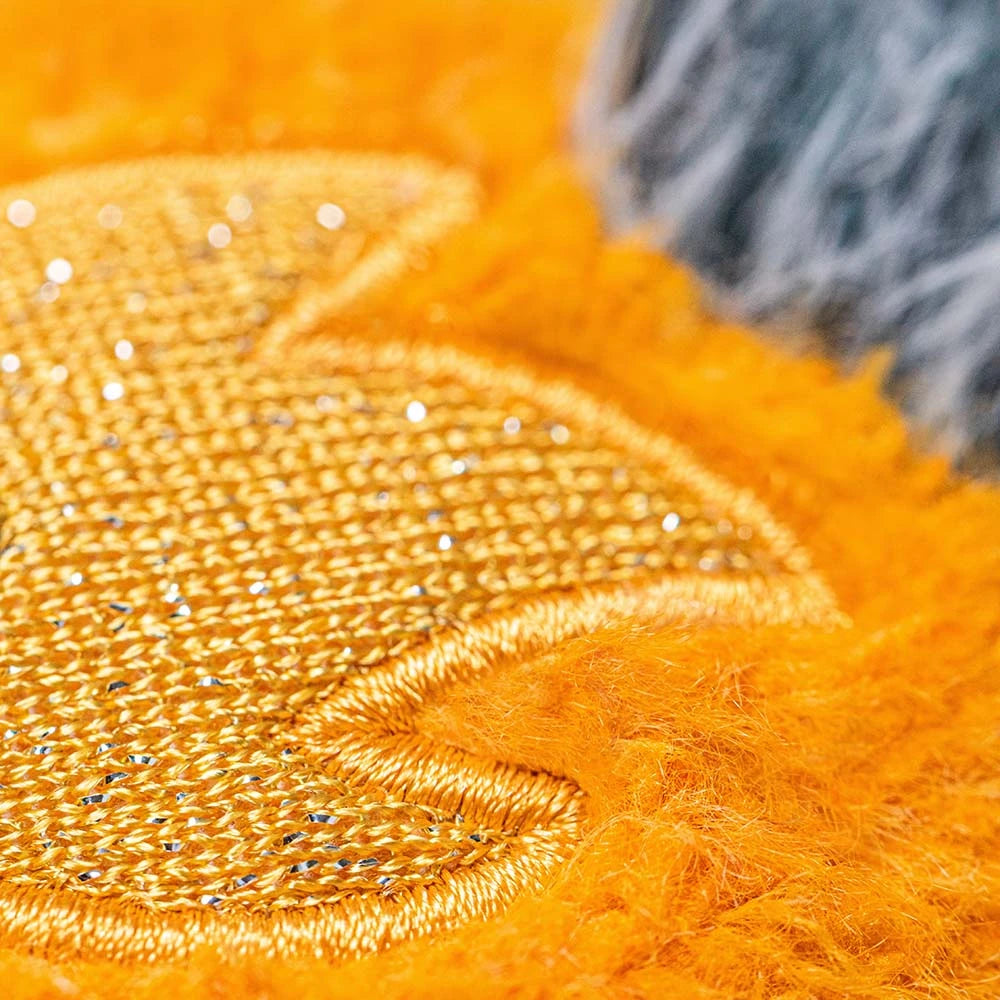 Close-up of the Jellycat Amuseables Space Comet plush toy, featuring textured yellow fabric with gold embroidery and orange fur, next to a patch of gray and black fuzzy material.