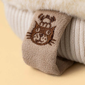 Close-up of a beige fabric tag with a brown embroidered cat face on a cream ribbed textile from Jellycat’s Amuseables Fitswell Mitten.