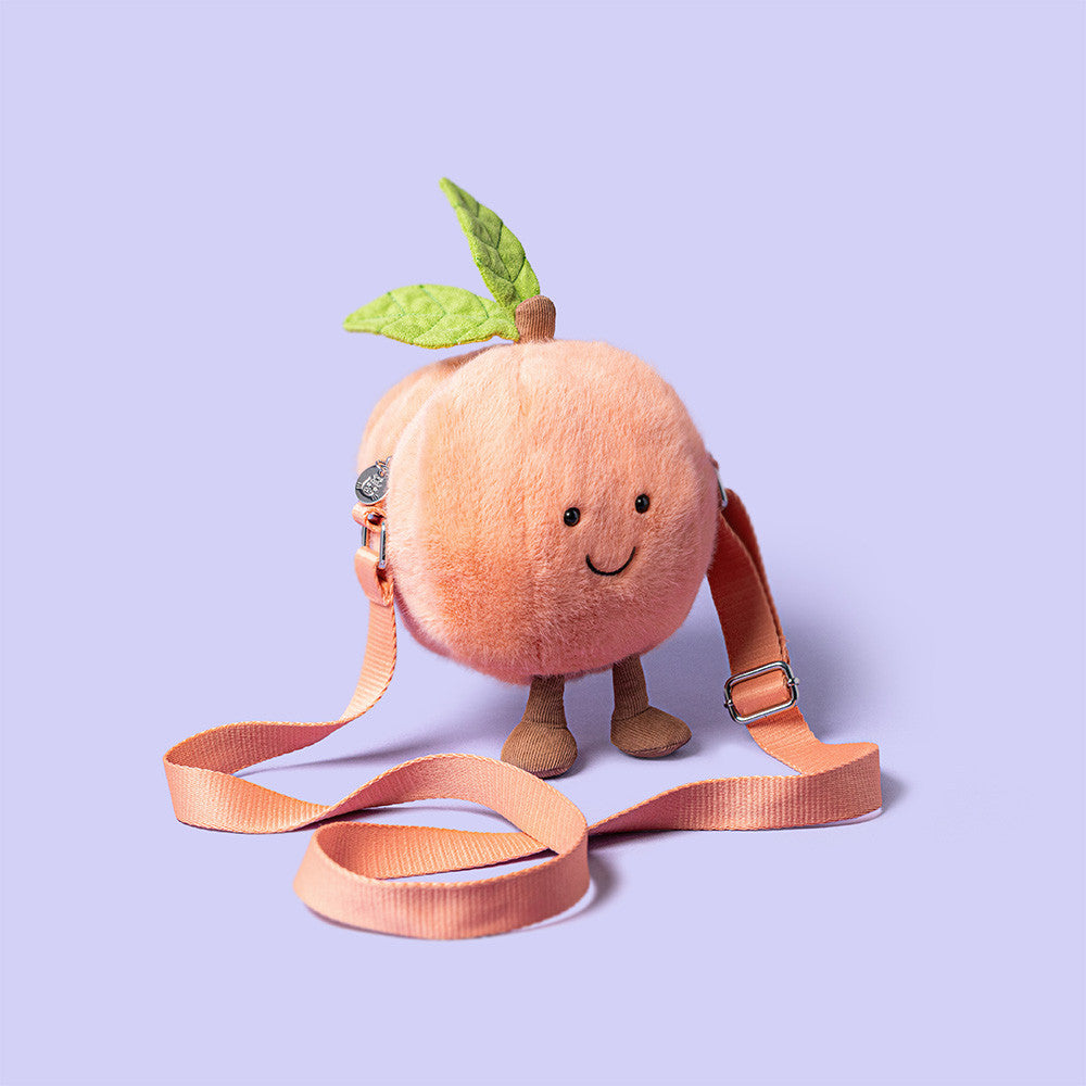 The Jellycat Amuseables Peach Shoulder Bag features a smiling plush peach with a green leaf, small legs, and peach-colored strap, shown on a light purple background.