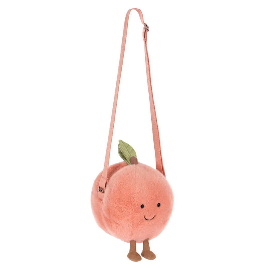 The Jellycat Amuseables Peach Shoulder Bag features a long pink strap, green felt leaf, small legs, and a smiling face.