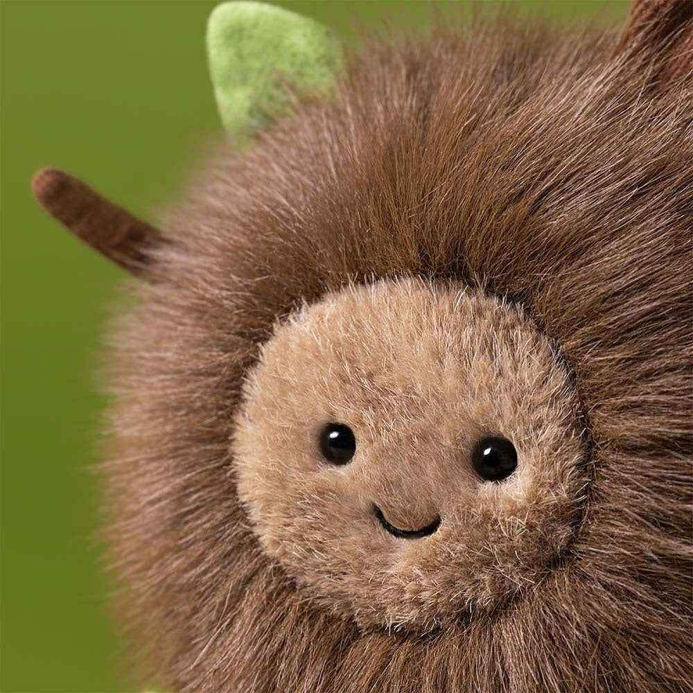 Close-up of Jellycat's Amuseables Mulshi Woodland Floor plush, featuring a round, furry brown body, beige face, black button eyes, stitched smile, and green and brown leaf-like ears against a green background.