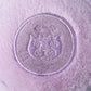 A close-up of a round, embroidered purple patch with a stylized cat face on soft lavender fabric, inspired by the Jellycat Amuseables May Macaron - Lavender plush toy.
