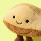 The Jellycat Amuseables Margaux Madeleine is a plush toy shaped like a pastry with a smiling face, black eyes, and brown legs, displayed on a light yellow background.