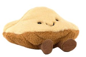 The Jellycat Amuseables Margaux Madeleine is a plush toy shaped like a madeleine with a smiling face and small brown corduroy legs, shown against a white background.