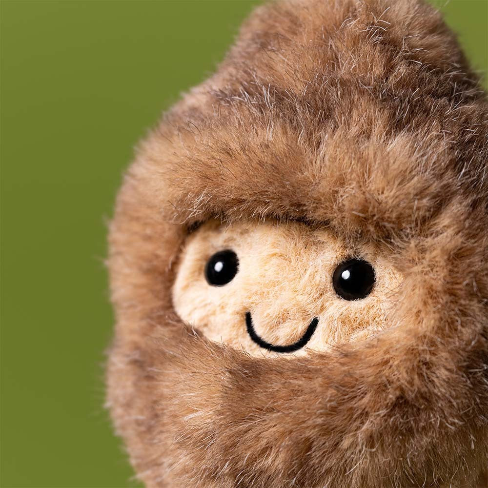 A close-up of the Jellycat Amuseables Peanut Yeti Outfit plush, featuring brown fur, black button eyes, and a stitched smile against a plain green background.