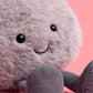 A close-up of Jellycat's Amuseables Pebble, a dependable plush toy with a fuzzy round gray body, black button eyes, a stitched smile, and corduroy legs, set against a pink background.