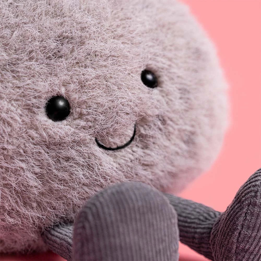 A close-up of Jellycat's Amuseables Pebble, a dependable plush toy with a fuzzy round gray body, black button eyes, a stitched smile, and corduroy legs, set against a pink background.