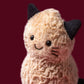 A Jellycat Amuseables Peanut ‘Cat’ plush toy, featuring beige fur, black ears, and embroidered facial details, sits against a solid maroon background.