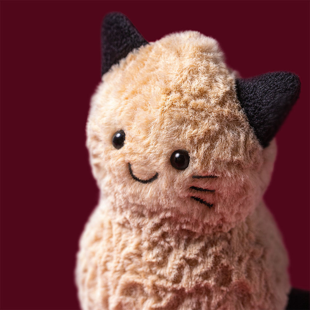 A Jellycat Amuseables Peanut ‘Cat’ plush toy, featuring beige fur, black ears, and embroidered facial details, sits against a solid maroon background.