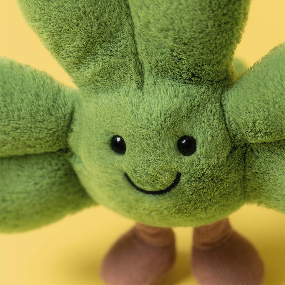The Jellycat Amuseables Siofra Shamrock is a green leafy plush with a smiling face, black eyes, and brown feet, shown on a vibrant yellow background.