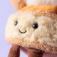 Close-up of a Jellycat Amuseables Mirielle St. Honoré Cake plush, featuring beige and brown fur, black button eyes, a stitched smile, and two brown corduroy legs against a light purple background.