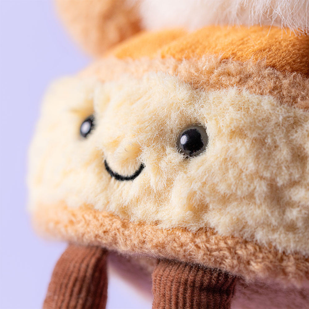 Close-up of a Jellycat Amuseables Mirielle St. Honoré Cake plush, featuring beige and brown fur, black button eyes, a stitched smile, and two brown corduroy legs against a light purple background.