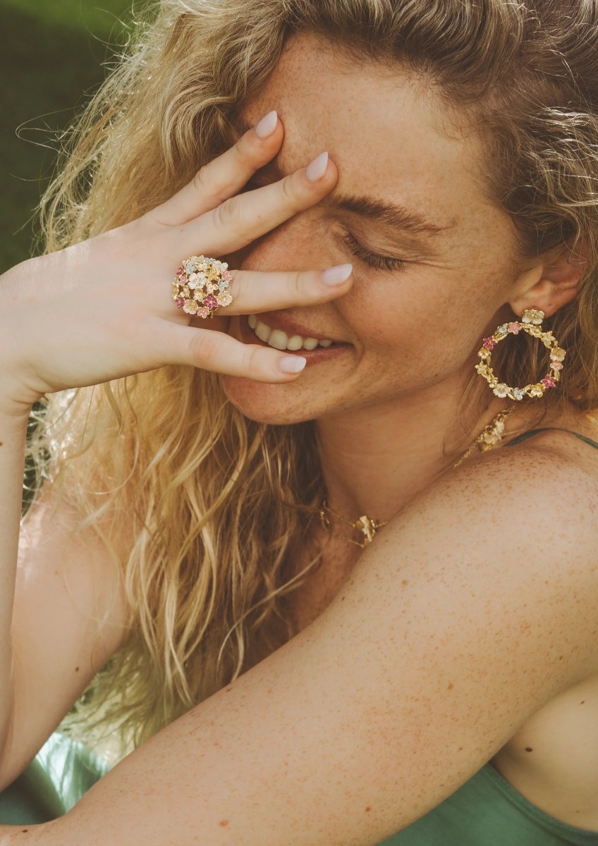 A woman with long curly blonde hair smiles with her eyes closed, resting her hand on her face. She wears floral jewelry by Anabel Aram, including the Wildflowers Gathered Ring and matching earrings and necklace.