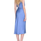 A woman with long blonde hair stands with her back to the camera against a white background, wearing the Anine Bing Bar Silk Skirt in light blue as a midi-length dress paired with black heeled sandals.