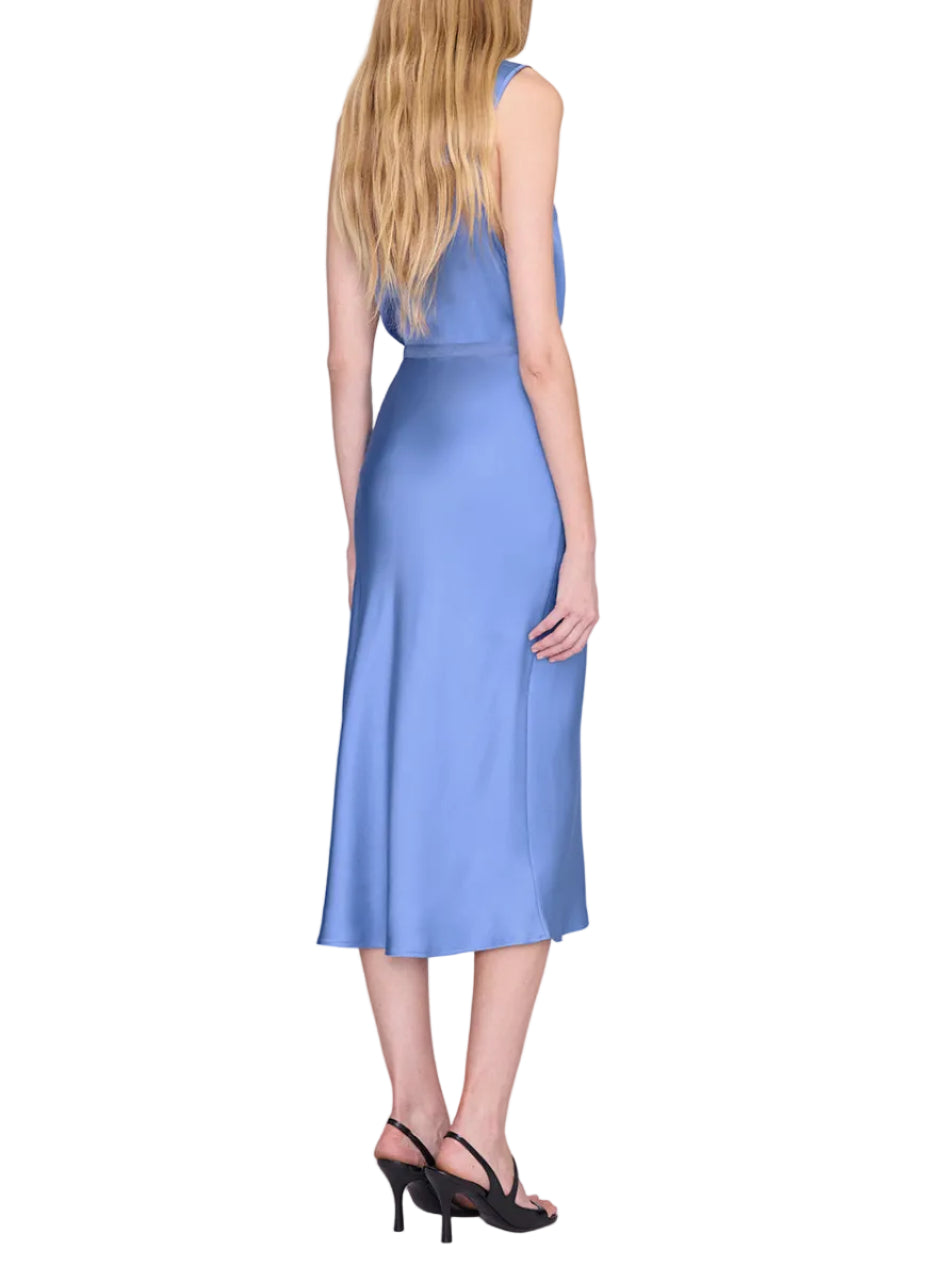 A woman with long blonde hair stands with her back to the camera against a white background, wearing the Anine Bing Bar Silk Skirt in light blue as a midi-length dress paired with black heeled sandals.