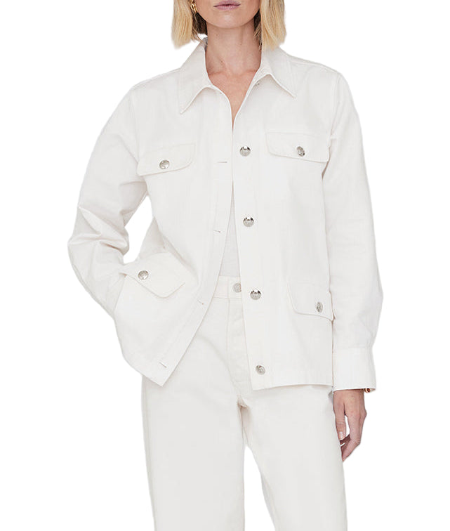 A woman wears the Corey Jacket by Anine Bing, a white cotton canvas jacket with flap pockets, paired with white pants and standing against a plain background.