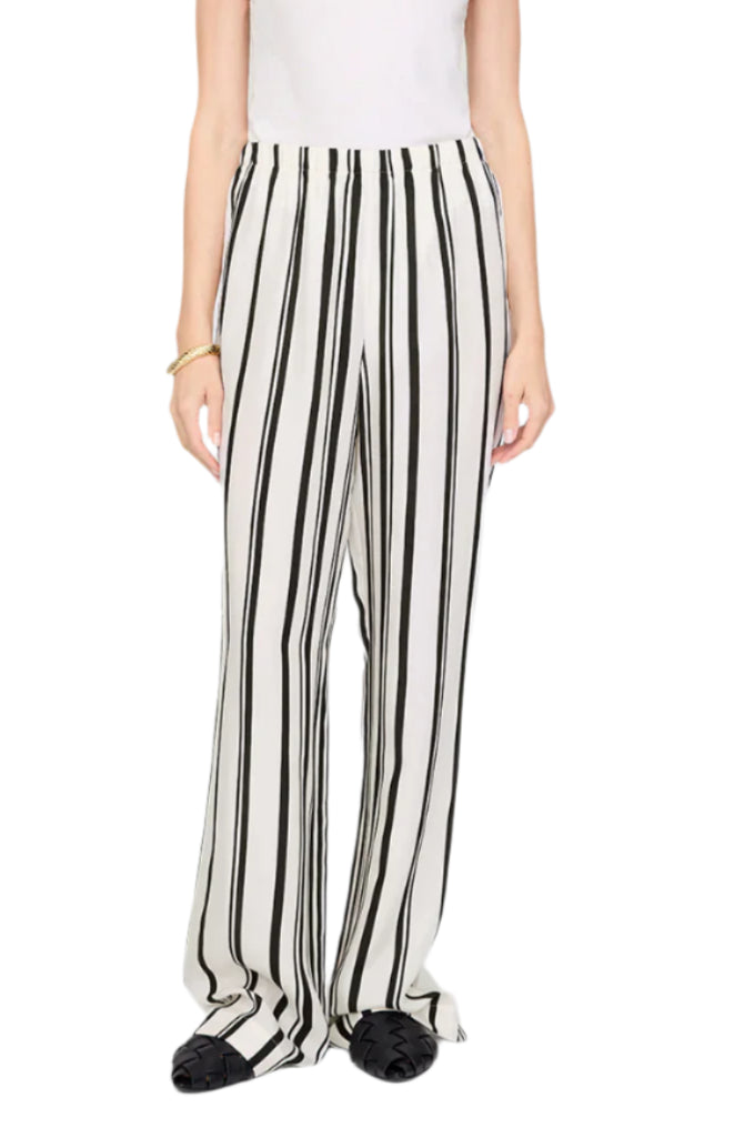 A person wears the Elain Pant Monogram by Anine Bing in an ivory and black stripe cupro blend, styled with a white top, black shoes, and a gold bracelet.