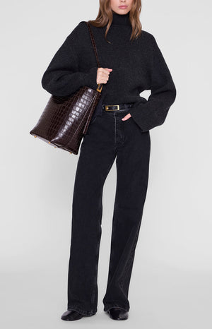 A person wears a dark sweater and black jeans while carrying an Anine Bing Elly Bucket Embossed Bag, a dark brown, croc-embossed leather bucket bag with gold-tone hardware, over their shoulder.