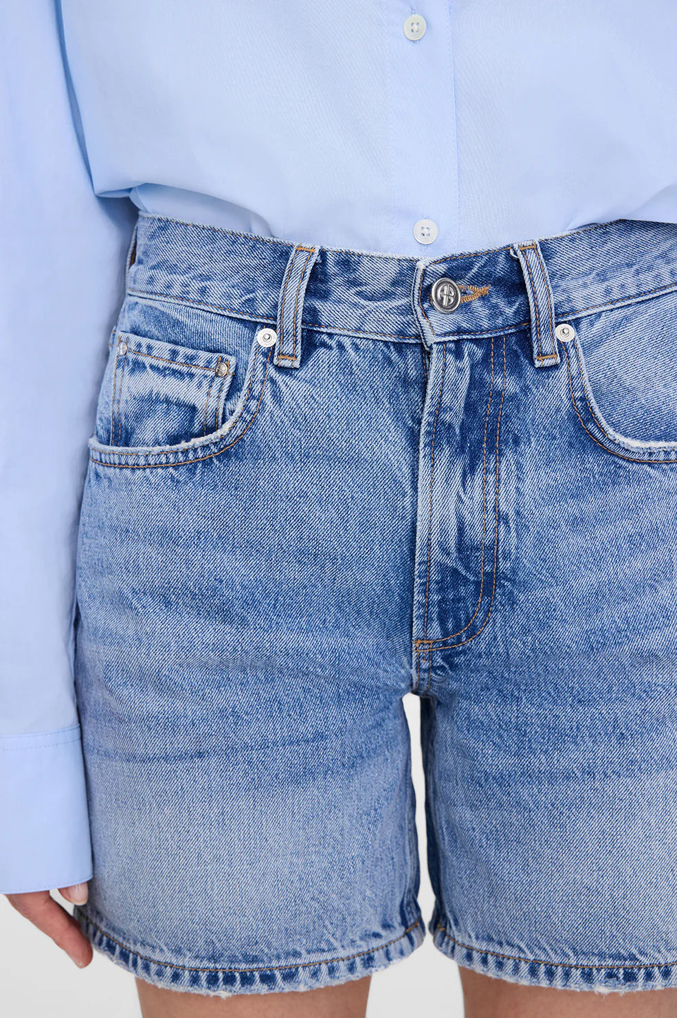 A person wears the Anine Bing Heath Short—high-waisted, vintage blue wash baggy shorts—paired with a light blue button-up shirt tucked in. Only the torso and upper legs are shown.