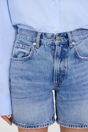 A person wears the Anine Bing Heath Short—high-waisted, vintage blue wash baggy shorts—paired with a light blue button-up shirt tucked in. Only the torso and upper legs are shown.