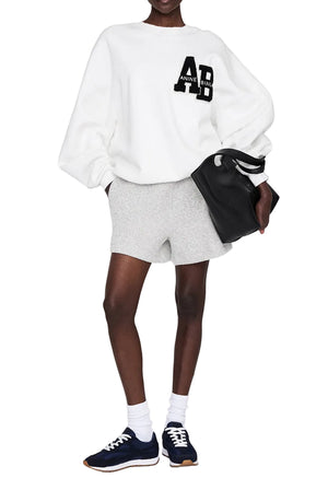 A person wearing a white oversized Anine Bing Kam Short Framed Monogram sweatshirt, gray shorts, navy sneakers, white socks, sunglasses, and holding a black tote bag stands against a plain background.
