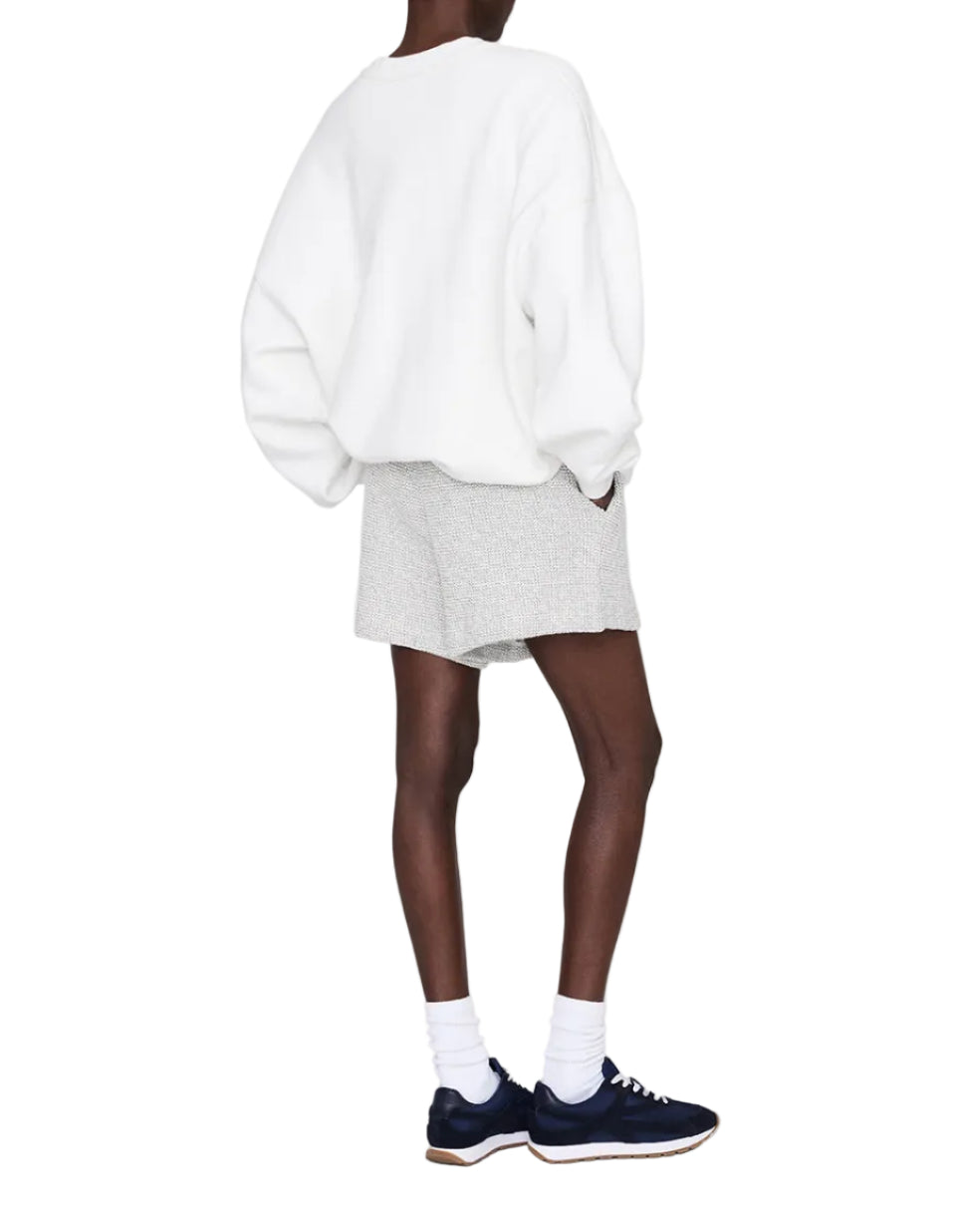 A person faces away from the camera in an off-duty essential look: wearing a white oversized sweatshirt and Anine Bing's Kam Short Framed Monogram gray shorts, with white socks and navy sneakers against a plain background.