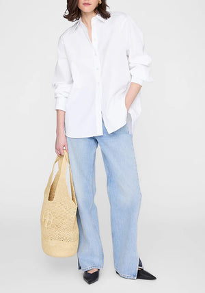 A person wearing a white button-up shirt, light blue jeans, and black pointed shoes holds the Anine Bing Leah Logo Hobo bag, standing against a plain background.