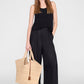 A woman stands facing forward against a plain light background, wearing a black sleeveless top, loose black pants, and sandals, holding the Anine Bing Lili Raffia Tote in her left hand.