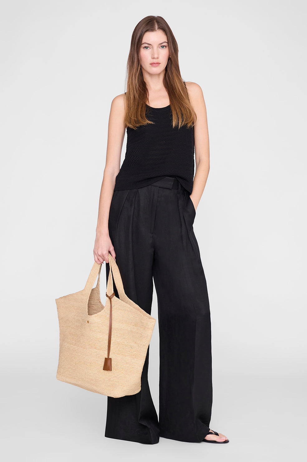 A woman stands facing forward against a plain light background, wearing a black sleeveless top, loose black pants, and sandals, holding the Anine Bing Lili Raffia Tote in her left hand.