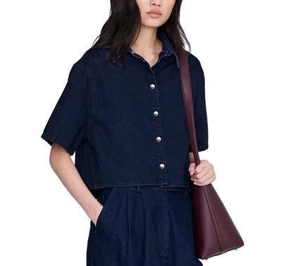 A woman wearing the Anine Bing Lincoln Shirt, a cropped denim top in an ink-blue wash, with matching pants holds a maroon shoulder bag.