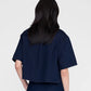 A person with long dark hair is shown from the back, wearing the Anine Bing Lincoln Shirt—a cropped denim top—in an ink-blue wash with matching pants against a plain light background.