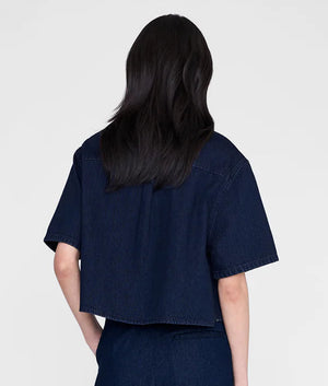 A person with long dark hair is shown from the back, wearing the Anine Bing Lincoln Shirt—a cropped denim top—in an ink-blue wash with matching pants against a plain light background.