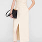 A woman wearing the Anine Bing Tyla Top, styled as a sculpted beige strapless piece and paired with a beige midi skirt featuring a front slit, black heeled sandals, and a black clutch bag, stands against a plain background.