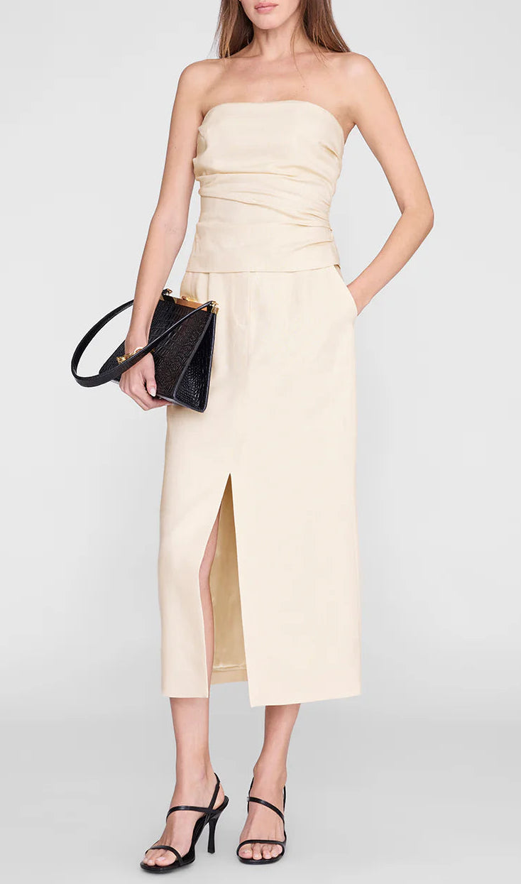 A woman wearing the Anine Bing Tyla Top, styled as a sculpted beige strapless piece and paired with a beige midi skirt featuring a front slit, black heeled sandals, and a black clutch bag, stands against a plain background.