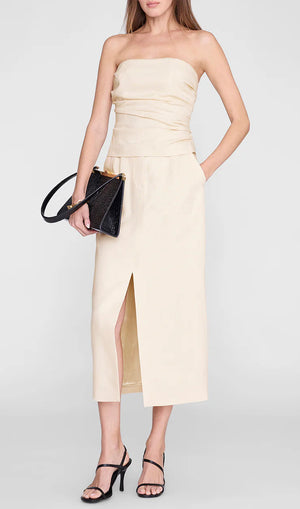 A woman wearing the Anine Bing Tyla Top, styled as a sculpted beige strapless piece and paired with a beige midi skirt featuring a front slit, black heeled sandals, and a black clutch bag, stands against a plain background.