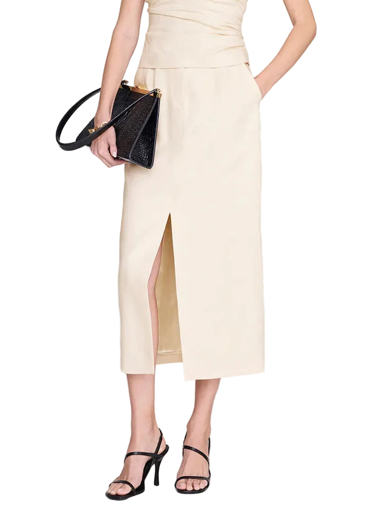A woman styles the Anine Bing Meredith Skirt—a cream, high-rise tailored midi with a front slit—with black strappy heels and a matching black handbag. Her partially visible top adds to the skirt’s versatility, making it ideal for both office and after-hours looks.
