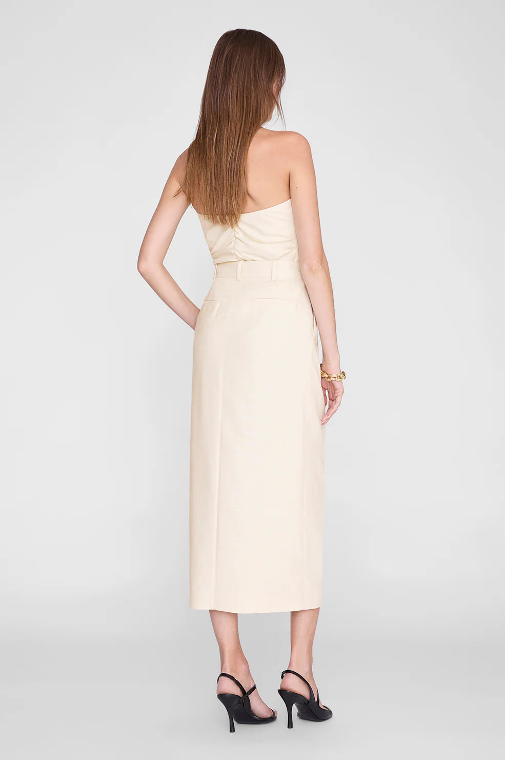 A woman stands facing away, wearing the Tyla Top by Anine Bing styled as a versatile beige strapless midi dress with a belt that accentuates her sculpted shape, paired with black high-heeled shoes against a plain light background.