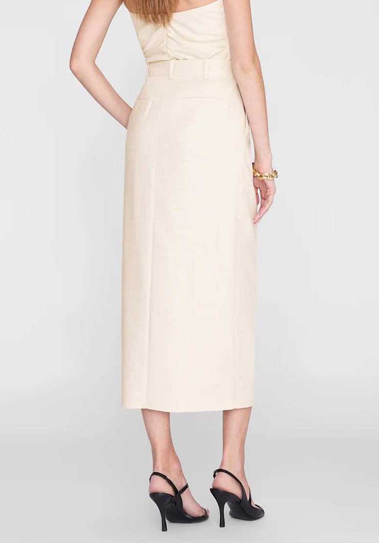 A woman stands facing away against a plain background, wearing the Anine Bing Meredith Skirt in beige—a high-rise waist midi skirt—paired with a matching top, black heeled sandals, and a gold bracelet.