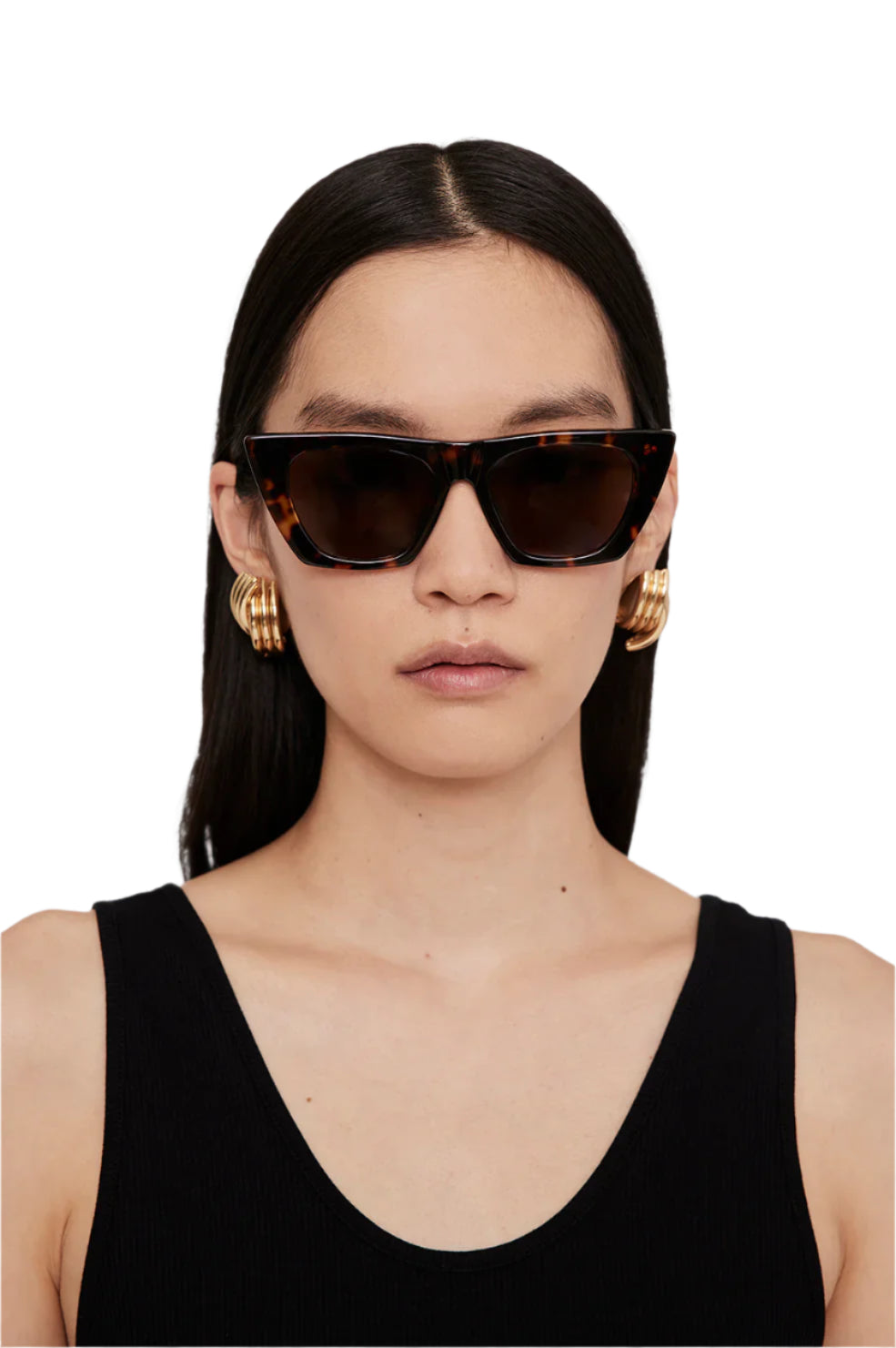A woman with straight dark hair wears Anine Bing Paris Sunglasses—cat-eye tortoise acetate frames with polarized lenses—plus gold hoop earrings and a sleeveless black top against a plain background.