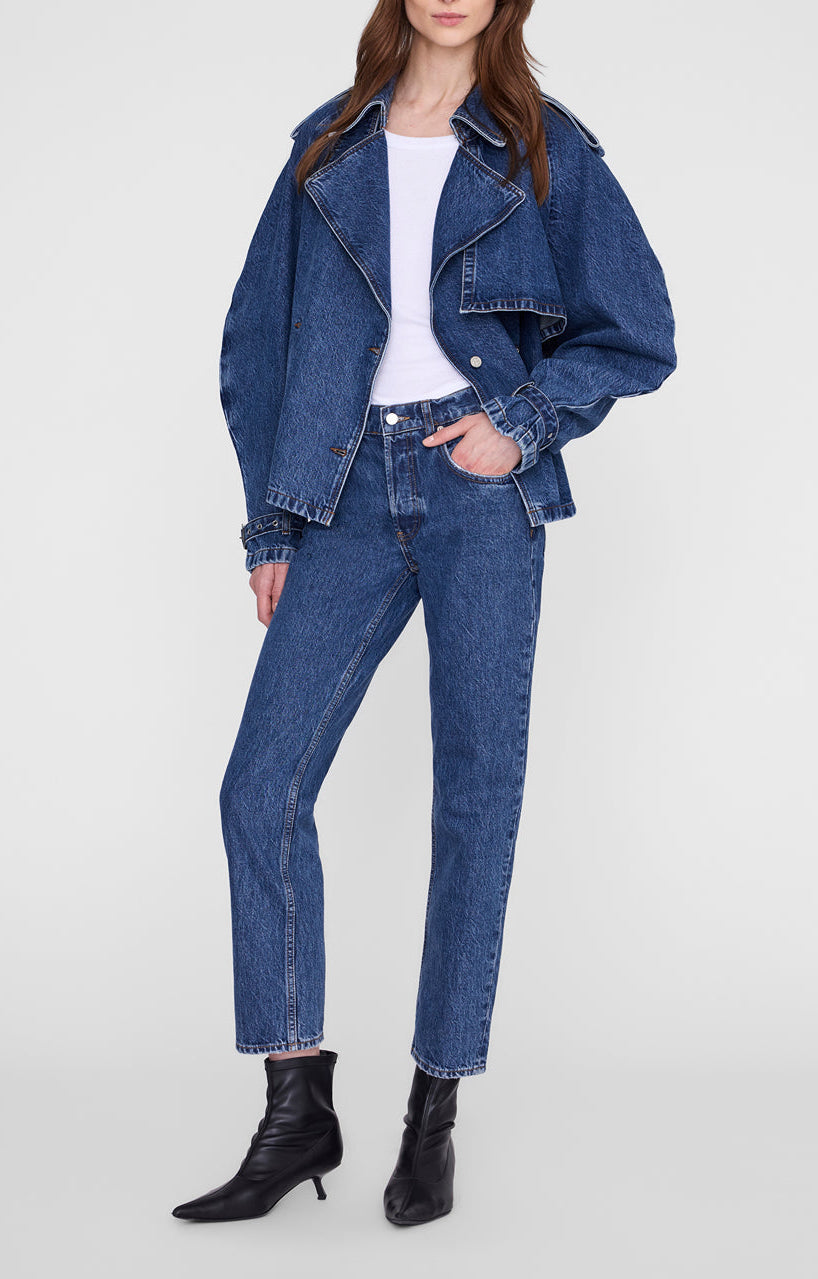 A woman stands against a plain light background wearing the Anine Bing Pascale Trench as transitional outerwear, paired with high-waisted blue jeans, a white top, and black ankle boots.