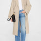 Person wearing the Anine Bing Rickie Maxi Trench in beige cotton-twill, styled with a white t-shirt, blue jeans, black shoes, and a black handbag, standing against a plain light background.