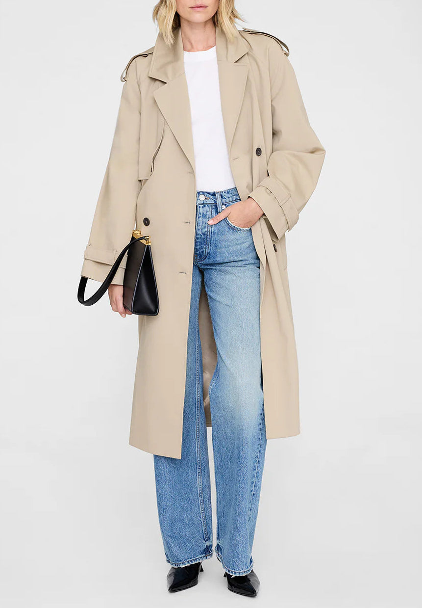 Person wearing the Anine Bing Rickie Maxi Trench in beige cotton-twill, styled with a white t-shirt, blue jeans, black shoes, and a black handbag, standing against a plain light background.