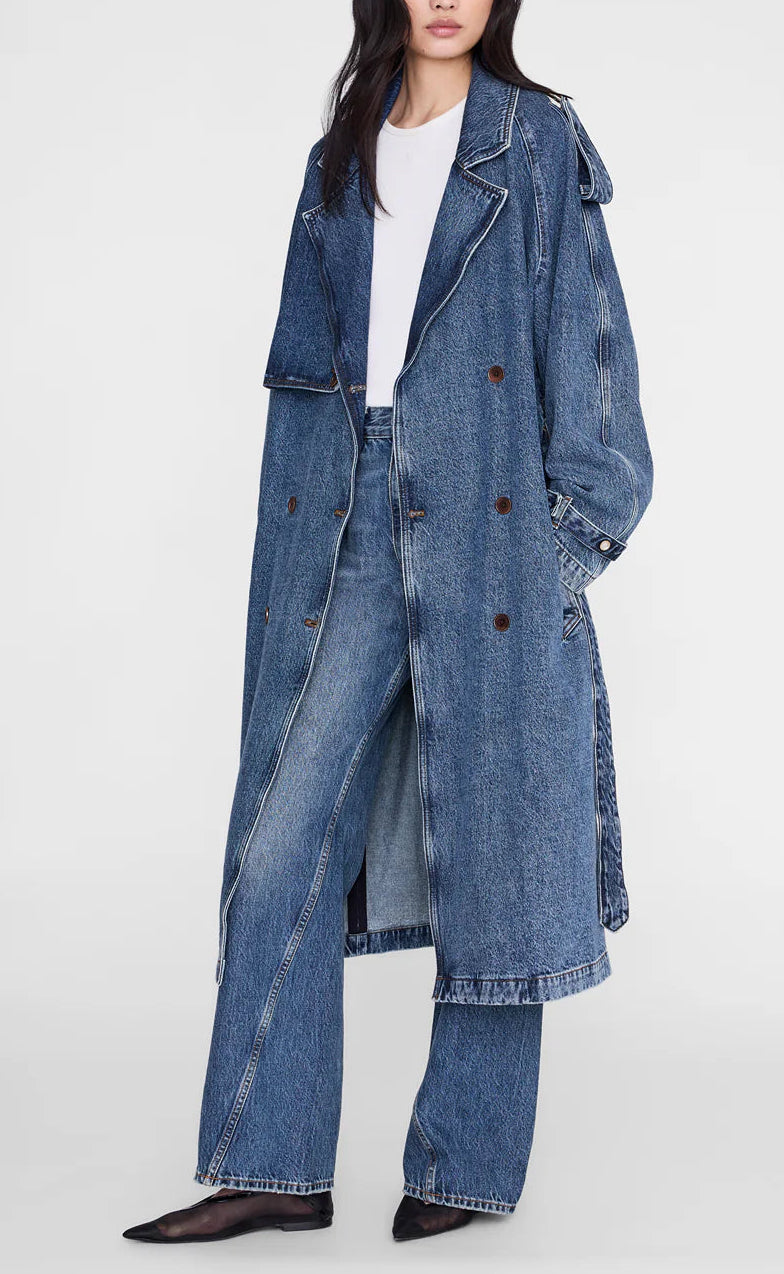 A woman wears the Rickie Maxi Denim Trench by Anine Bing over matching denim pants, a white top, and pointed black shoes for a chic, menswear-inspired look.