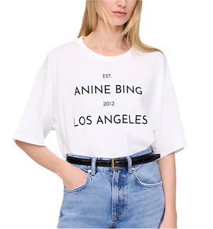 A woman wears the Rudy Tee by Anine Bing—a white t-shirt featuring "EST. ANINE BING 2012 LOS ANGELES"—styled with light blue jeans and a black belt for a relaxed, branded look.